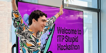 Lachlan raising their arms energetically in front of a pink TV that says Welcome to ITP Stupid Hackathon