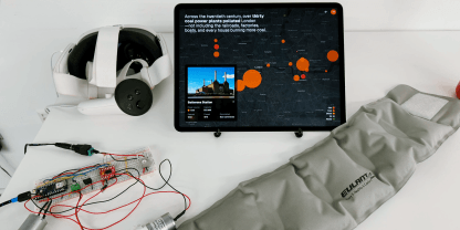 Grey neck pillow outstretched on a table with a breadboard and air pumps alongside a Meta Quest 3 VR headset and iPad showing a map of London